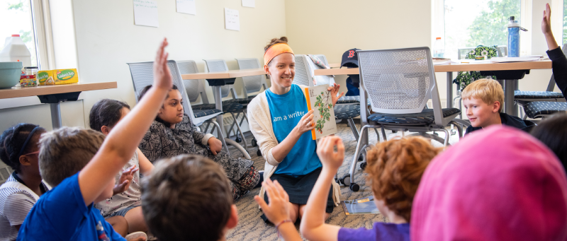 A teacher reading and displaying a picture book while a crowd of children raise their hands enthusiastically.